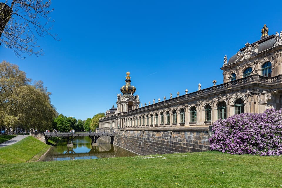 The Zwinger's Crown Gate and long gallery above a grassy moat, with a flowering lilac bush in the foreground under a clear blue sky.